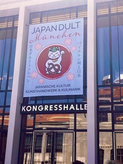 Entrance of the Japandult fair in Munich. The glass front of a modern business building sports a large Banner. The logo on it is a cartoon cat with a pretzel on a red circle.