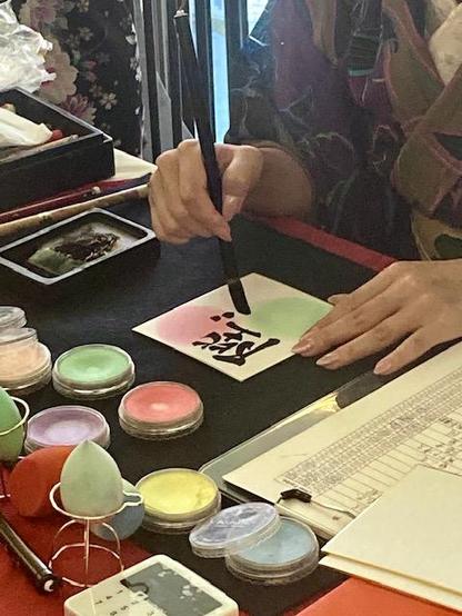 Closeup of the desk of a female calligrapher. She is currently finishing a name written in Kanji. There are pots with colorful powder and sponges on her desk that she used to add a dash of pink and green color to the piece of paper she is writing on.