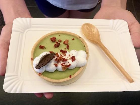 Top down photo of a small Japanese cake and a spoon. The cake is round and approximately palm-sized. It is bright green due to the Macha flavor and is topped with cream, a piece of chocolate and pieces of dried strawberries. The cake is placed on a tiny cardboard tray held by a person but you only see parts of their hands.