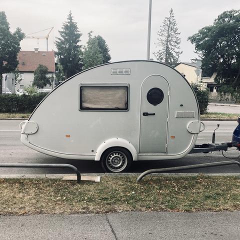 A small metal trailer parked on the side of the road. It has a very distinct tear drop shape with a single axle, a small door and a tiny window. The car pulling it is out of frame.