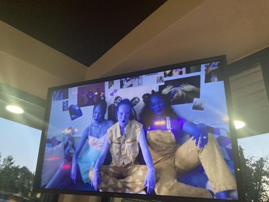 A TV in a diner showing  3 women sitting on a sofa. Their skin tones are blue due to some signal or wiring problem. A colorful contrast to their yellow pants.